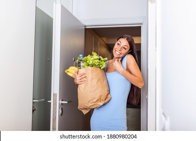 Pretty Woman Arriving Home Carrying Groceries In A Paper Bag Opening The Door And On A Phone Call - Multi Tasking. Young House Wife With Groceries Arriving Home