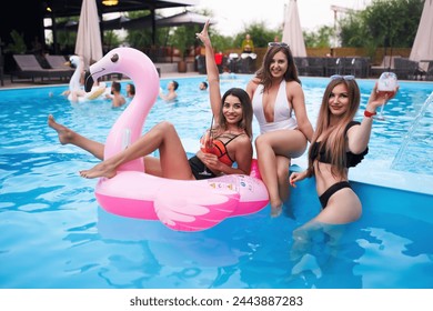 Pretty woman in bikini swimwear hanging out on inflatable pink flamingo mattress on night pool party. Friends partying with cocktails in resort villa swimming pool. Girls toast, cheer with drinks. - Powered by Shutterstock