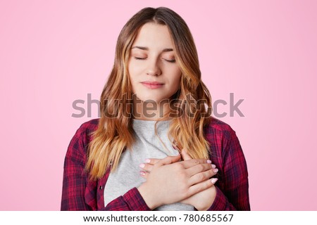 Pretty woman with closed eyes, keeps hands on chest, expresses her sympathy to someone, being thankful to close friend, isolated over pink background. Cute adorable grateful young female indoor