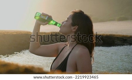 Pretty woman drinking beer from green glass bottle as she is standing inside a reef pool with ocean water at sunset. Enjoying cold alcoholic drink on seaside in tropical paradise. Soft focus. 