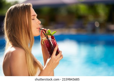 Pretty woman drinks refreshing cocktail decorated with mint, cherry and lemon. Female enjoys a drink near swimming pool with blue water. Girl chilling with beverage in tropical sun. Vacation concept - Shutterstock ID 2159843685