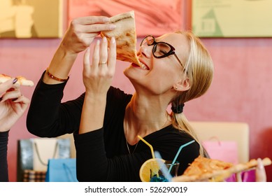 Pretty woman eating pizza. Girl wearing glasses, golden ring and bracelet on her hand. She has ponytail. Enjoying the meal. Italian restaurant with colorful interier.