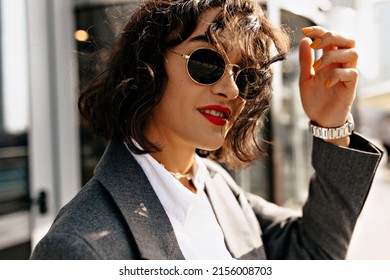 Pretty Woman In Grey Jacket And White Shirt In Sunglasses With Red Lipstick Is Touching Hair And Posing Near Her House. Beautiful Lady In Grey Oversize Jacket And Pants Walks Outside