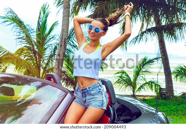 Pretty woman have fun at luxury car in\
vacation, posing near sea side with palm trees, wearing hight\
waisted shorts and stylish round\
sunglasses.