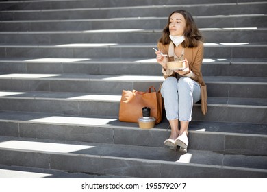 Pretty Woman Have Outdoor Lunch Near Office Building While Sitting On The Stairs. Healthy Meal For Takeaway. With Lowered Protective Mask On Her Chin And Social Distance Alone. Copy Space.