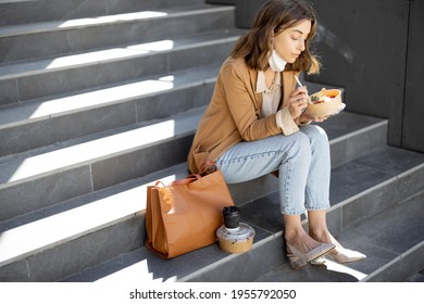 Pretty Woman Have Outdoor Lunch Near Office Building While Sitting On The Stairs. Healthy Meal For Takeaway. With Lowered Protective Mask On Her Chin And Social Distance. Copy Space.