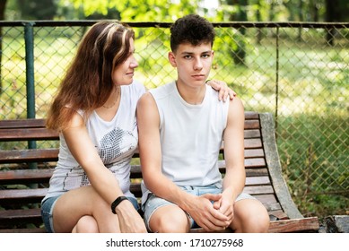 Pretty Woman With Her Teenage Son On A Bench In The Summer Sitting And Trying To Communicate. Parents And Teens Relationship Concept. Mom Hugs Her Son And Shows That She Loves Him