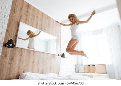 Pretty Woman Jumping On Bed In Bedroom With Mirror On The Wall And Window On Background