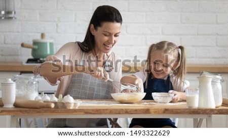 Pretty woman and little adorable daughter sitting at table in kitchen cook together stirring eggs for pie, cookies or pancakes, teach kid recipes, embodiment of ideal wife of mom and housewife concept