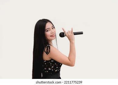 A Pretty Woman With Long Black Hair Holds A Microphone While Showing A Rock And Roll Hand Pose As She Beams For The Camera On Side View. Studio Shot Isolated On A White Background.