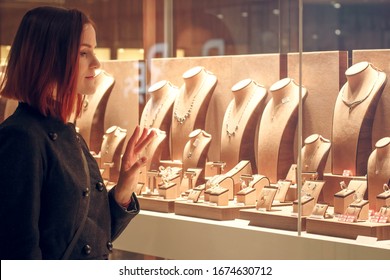 Pretty woman looking at jewelry in store window. Customer near jewellery. Dreamy red hair girl in classic style chooses silver, gold, diamonds, precious stones. Shopper - Powered by Shutterstock