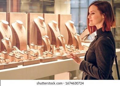 Pretty woman looking at jewelry in store window. Customer near jewellery. Dreamy red hair girl in classic style chooses silver, gold, diamonds, precious stones. Shopper - Powered by Shutterstock