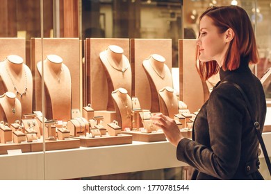 Pretty woman looking at jewelry in store window. Customer near jewellery. Dreamy red hair girl chooses silver, gold, diamonds, precious stones. Purchaser - Shutterstock ID 1770781544