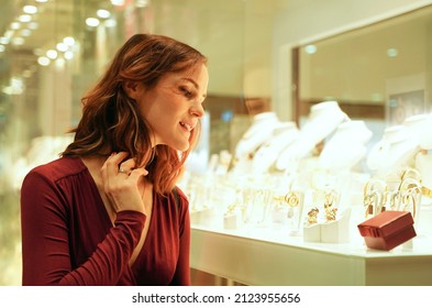 Pretty Woman Looking At Jewelry In Store Window. Interested Customer Near Jewellery. Girl Chooses Gold, Diamonds Or Precious Stones. Purchaser