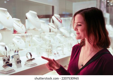 Pretty Woman Looking At Jewelry In Store Window. Girl Near Jewellery. Interested Customer Chooses Gold, Diamonds Or Precious Stones. Purchaser