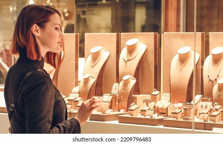 Pretty woman looking at jewelry in store window. Customer near jewellery. Dreamy red hair girl chooses silver, gold, diamonds, precious stones. Purchaser - Shutterstock ID 2209796483
