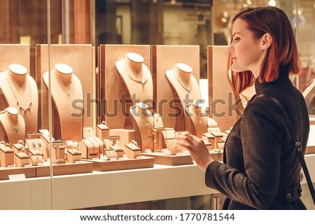Pretty woman looking at jewelry in store window. Customer near jewellery. Dreamy red hair girl chooses silver, gold, diamonds, precious stones. Purchaser