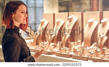 Pretty woman looking at jewelry in store window. Customer near jewellery. Dreamy red hair girl chooses silver, gold, diamonds, precious stones. Purchaser