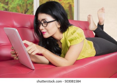 Pretty Woman Lying On The Red Couch While Playing With Tablet In Living Room