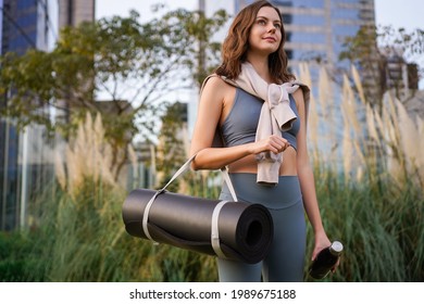 Hübsche Frau, die mit sportlicher Ausrüstung auf dem Stadtpark posiert, einen stilvollen Yogaanzug und einen Hufeisen trägt.