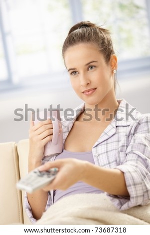 Pretty woman in pyjama sitting on couch with coffee mug, watching television in morning, changing channel with remote control.?