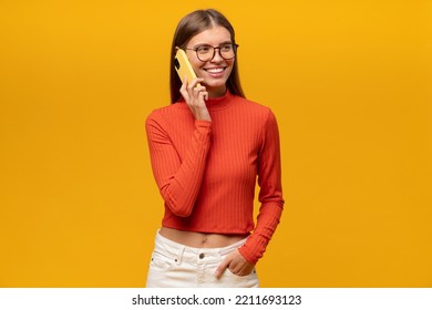 Pretty Woman In Red Turtleneck Talking On Phone With Her Friend, Sharing News, Plans, Gossips Putting Hand In Pocket On Yellow Studio Background. Mobile Network Connection