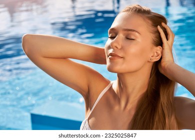 Pretty woman relaxing and sun bathing near swimming pool at luxury spa. Beautiful serene young female model on holiday travel resort enjoying vacation. Sun protection concept. Wellness beach club.