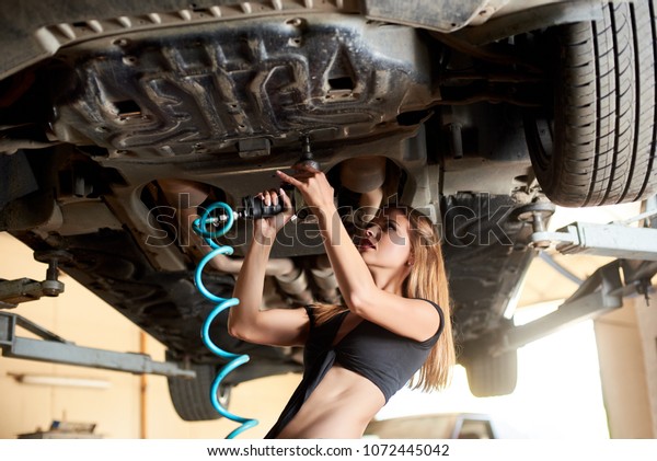 Pretty woman repairing the car with pneumatic key on\
hydraulic lift in a car repair shop. A girl in short T-shirt has a\
beautiful body