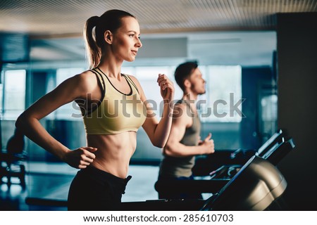 Pretty woman running on treadmill with fit young man on background