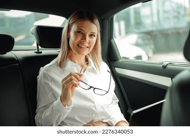 Pretty woman sales manager working on laptop sitting car backseat and looking at camera