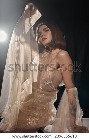 pretty woman with short hair posing in transparent dress posing near bathtub with black backdrop