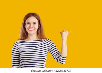 Pretty woman showing something isolated on solid yellow copy space background. Studio portrait of happy beautiful young lady in casual striped top smiling and pointing her thumb to copyspace side - Shutterstock ID 2206739719