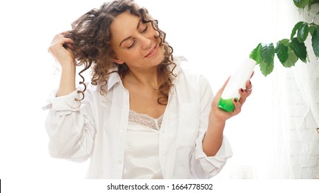 Pretty Woman Showing White Cosmetic Tube For Curly Hair Care In Her Hand At Home, White Room With Green Plants