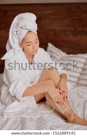 Pretty woman sitting on bed, spending time by taking care about perfect slim body after bath procedures, having white soft towel on head, putting wax strips on slender leg, beautification