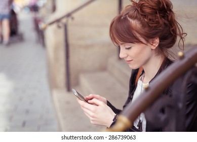 Pretty woman sitting on steps in an urban street to read an sms on her mobile phone, profile view