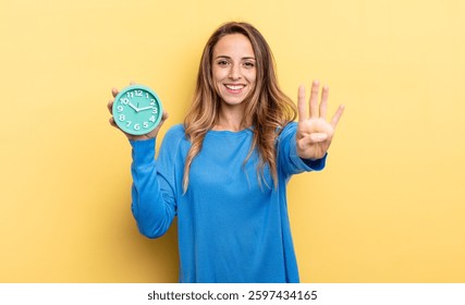 pretty woman smiling and looking friendly, showing number four holding an alarm clock
