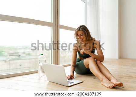 Pretty  woman in sportswear is sitting on the floor with  bottle of water and is using a laptop at home in the living room. Sport and recreation concept.
