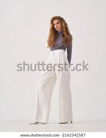 Pretty woman with stylish makeup and long flowing hair wearing sweater and loose trousers holds hand on hip on white background