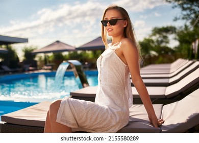 Pretty Woman Sunbathing And Chilling Near Swimming Pool On Beach Bed At Tropical Resort. Attractive Girl In White Dress And Sunglasses Lies In The Sun On Deck Chair In Spa, Relaxing On Hot Summer Day.