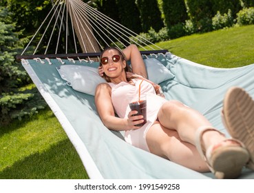 Pretty Woman Sunbathing In Hammock In Garden. Attractive Girl Relaxing On Vacation Smiling Having Drink. 