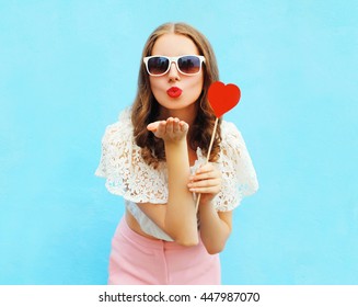 Pretty woman in sunglasses with red heart lollipop sends an air kiss over colorful blue background