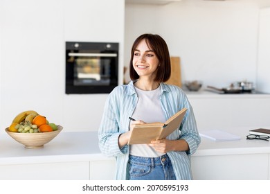 Pretty Woman Taking Notes, Holding Notepad And Pen, Standing In Kitchen Interior And Looking Aside, Dreaming And Smiling, Copy Space. Happy Young Lady Planning Her Day, Writing Tasks