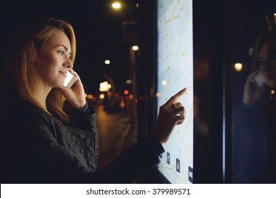 Pretty woman tourist talking on mobile phone while looks at map of the city on electronic bulletin board, young female calling with cell telephone while touching digital display of modern bus station