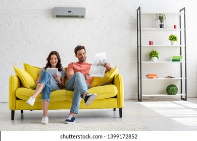 Pretty Woman Using Digital Table And Handsome Man Reading Newspaper While Sitting On Yellow Sofa Under Air Conditioner At Home