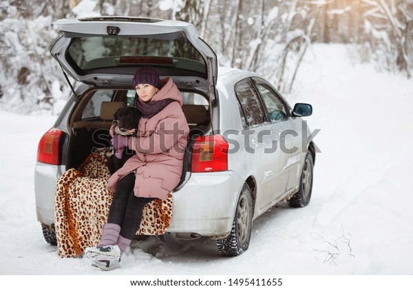 Pretty woman in warm jackets and hats and a funny\
black labrador retriever are sitting in the trunk of a car in the\
winter forest. Family happiness, care about nature. Purebred,  dog\
- man\'s friend.