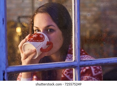 Pretty Woman In Warm Room Indoor At Home In Winter Night. Drinking Tea Looking Away. Candid Shot From Outdoor Trough The Window.