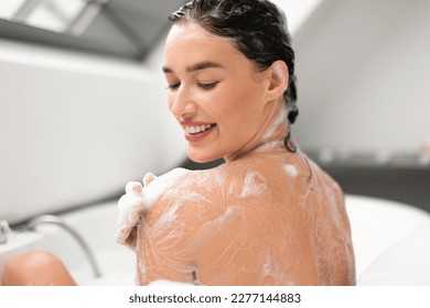 Pretty Woman Washing Body Taking Bath With Foam Applying Shower Gel On Shoulder Sitting Back To Camera In Bathtub In Modern Bathroom Indoors. Bodycare Beauty Routine Concept. Selective Focus
