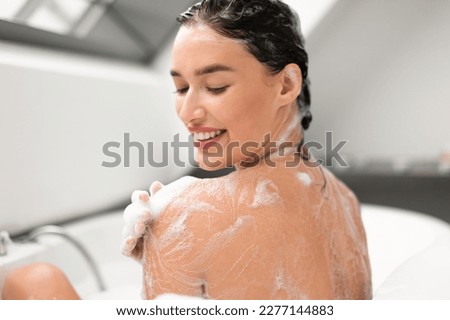 Pretty Woman Washing Body Taking Bath With Foam Applying Shower Gel On Shoulder Sitting Back To Camera In Bathtub In Modern Bathroom Indoors. Bodycare Beauty Routine Concept. Selective Focus