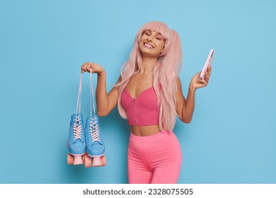 Pretty woman wearing Barbie style, posing on blue background in pink clothes with pink hair, holding roller-skates and phone in hands and smiles, Barbie fashion concept, copy space