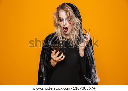 Pretty woman wearing black costume and halloween makeup holding smartphone isolated over yellow background